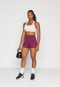 Sporu krūšturis gaiši rozā krāsā, augstu jostasvietu sarkanbrūni šorti, turot melnu kettlebellu. Baltas Nike zeķes un melnas sporta kurpes papildina apģērbu.