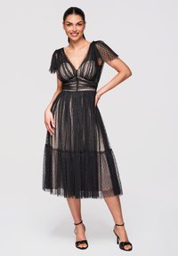 Femme debout, main sur la hanche, portant une robe midi noire transparente à pois et des sandales à talons hauts noires avec bride à la cheville, souriante à la caméra.