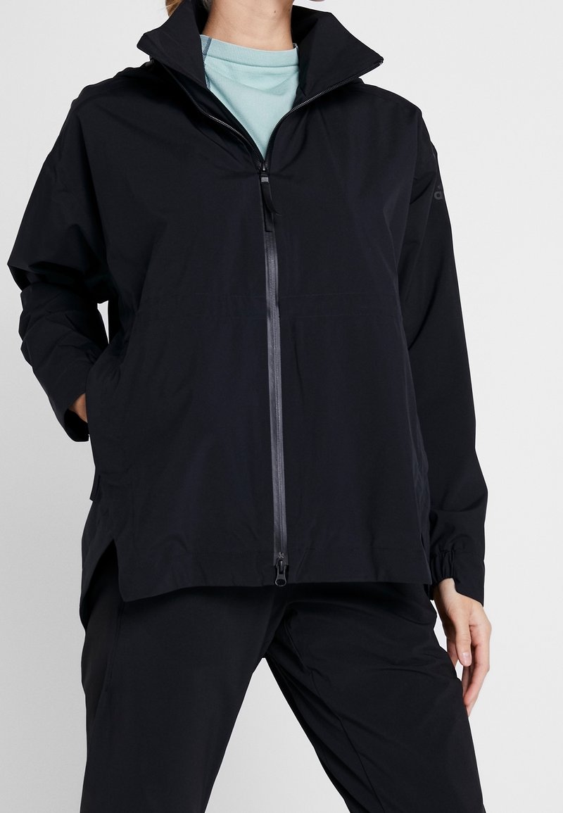 Personne portant une veste zippée noire par-dessus une chemise bleu clair, les mains partiellement dans les poches de la veste, debout devant un fond uni.