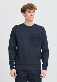Navy sweatshirt met lange mouwen, ronde hals en een voorzak; gemaakt van een gestructureerde stof. Gecombineerd met donkere jeans.