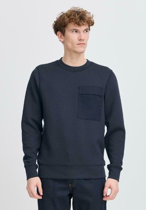 Navy sweatshirt with long sleeves, crew neck, and a front patch pocket; made from a textured fabric. Paired with dark jeans.