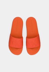 Paire de sandales slides orange vif avec de larges brides embossées de "CK" et des semelles texturées, présentées sur un fond clair uni.