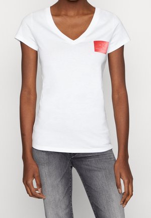 Femme portant un T-shirt blanc à col en V avec un petit patch logo rouge Armani Exchange sur la poitrine gauche, associé à un jean gris.