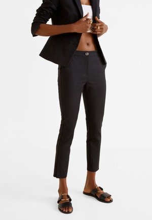 Person wearing cropped black pants, a black blazer over a white top, and black slide sandals with gold buckles, standing on a white background.