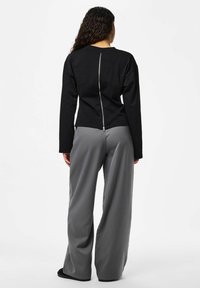 Haut noir à manches longues avec une fermeture éclair argentée visible dans le dos, associé à un pantalon large gris à rayures fines et des sandales noires.