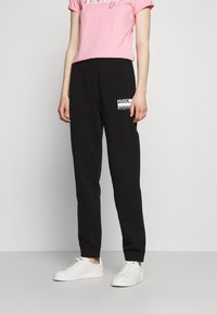 Schwarze Jogginghosen mit elastischen Bündchen und einem HUGO-Logo auf der rechten Tasche. Kombiniert mit einem rosa T-Shirt mit kurzen Ärmeln. Weiße Sneaker runden den Look ab.