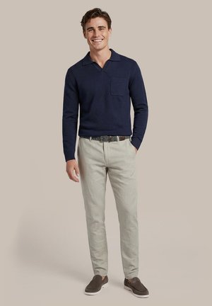 Man lacht, draagt een marineblauw gebreid shirt met lange mouwen en kraag, beige broek, geweven riem en bruine instapschoenen, staand tegen een effen achtergrond.