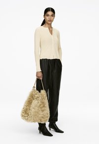 Hellbeige Langarm-Cardigan mit gewelltem Saum und Knöpfen, kombiniert mit schwarzen Satin-Hosen und einer flauschigen, beige farbenen Tragetasche. Schwarze Knöchelstiefel.