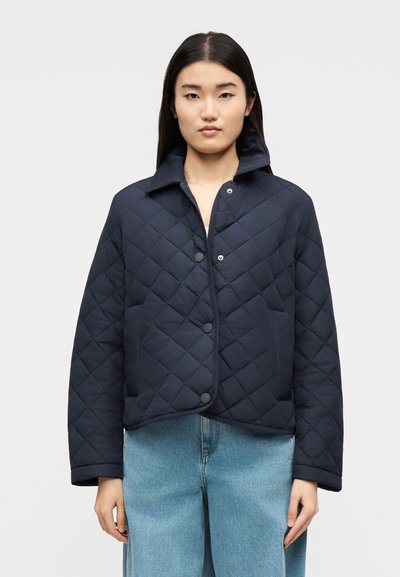 Jeune femme portant une veste matelassée bleu marine avec des boutons-pression et un jean large bleu clair, debout devant un fond uni.