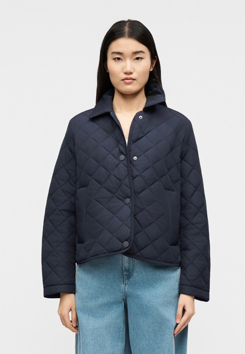 Νεαρή γυναίκα με σκούρο μπλε καπιτονέ μπουφάν με κουμπιά και ανοιχτό μπλε τζιν παντελόνι φαρδιάς γραμμής, που στέκεται μπροστά σε απλό φόντο.