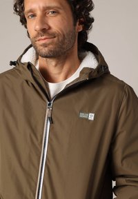 Bruine zip-up jas met een gevoerde capuchon, voorzien van een logopatch, zilveren rits en een gestructureerde binnenvoering. Glad materiaal met een praktische uitstraling.