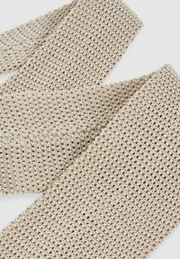 Beige színű szőtt textilöv, nyitott kötött mintával, puha tapintással és lekerekített élekkel. Különböző stílusopciókhoz megfelelő.