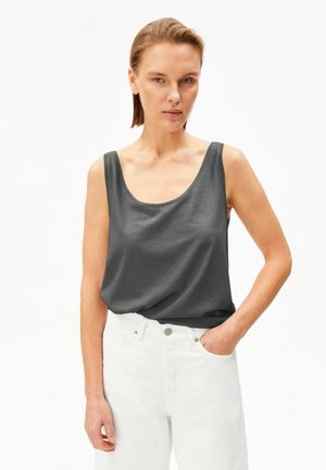 Donna con capelli corti che indossa una canottiera grigio scuro senza maniche infilata in pantaloni bianchi a vita alta, in piedi con una mano in tasca.