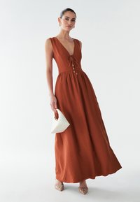 Maxi šaty v rustovej farbe s výstrihom do V a detailom na viazanie, so priliehavým korzetom a voľnou sukňou. Model drží bielu geometrickú kabelku.