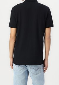 Person med en enfärgad svart kortärmad polo tröja och ljusblå jeans, sedd bakifrån mot en enfärgad bakgrund.