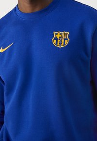Sudadera azul confeccionada con mezcla de algodón. Cuenta con el logo dorado de Nike en el pecho izquierdo y un emblema del club Barcelona en amarillo y azul. Cuello redondo.