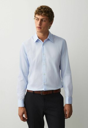 Young man wearing a light blue long-sleeve dress shirt, tucked into black trousers with a brown leather belt, standing against a plain wall.