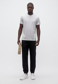 Άνδρας με γκρι αθλητική μπλούζα και μαύρο παντελόνι που κρατά μια μπεζ λωρίδα αντίστασης, στέκεται μπροστά σε λευκό φόντο.