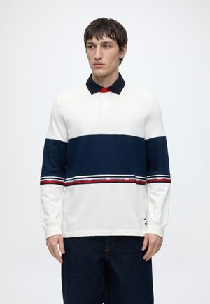 Giovane uomo che indossa una maglia da rugby bianca e blu navy a maniche lunghe con dettagli a righe rosse, bianche e blu e jeans scuri su uno sfondo semplice.