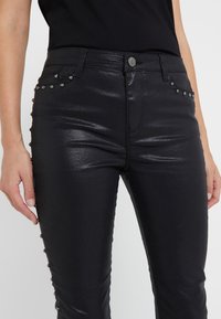 Pantaloni neri in finto cuoio con finitura lucida, caratterizzati da una vestibilità aderente, borchie decorative argento sui lati e un classico design a cinque tasche.