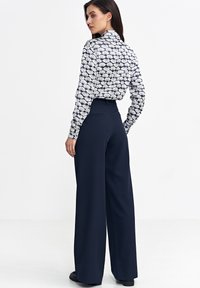Długie granatowe spodnie i dopasowana, biała bluzka z długim rękawem w granatowy wzór muszelkowy. Strój ma eleganckie detale i subtelne tekstury.