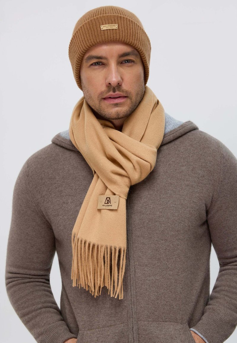 Brown knit beanie and a beige scarf with fringed ends, worn over a gray hoodie. Scarf features a small logo tag. Smooth texture.