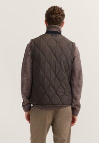 Braun gepolstertes Weste mit wellenförmigem Muster und hohem Kragen, kombiniert mit einem grob gestrickten Pullover. Das Outfit umfasst Hosen in neutralen Farbtönen.