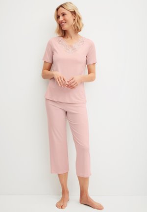 Vrouw staat blootsvoets, draagt een lichtroze shirt met korte mouwen en kanten halslijn en bijpassende cropped broek tegen een witte achtergrond.