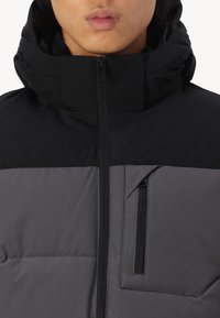 Puffervest med en sort øverste sektion og grå nederste sektion. Har en høj krave og en lynlåslommer på brystet med sort hardware.