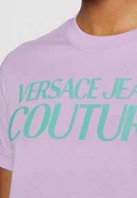 Närbild av en person som bär en lavendelfärgad t-shirt med grön text "Versace Jeans Couture" tryckt över bröstet.