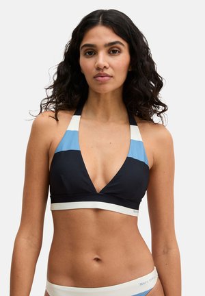 Frau mit gewellten dunklen Haaren, die ein Neckholder-Bikinitop mit schwarzen, blauen und weißen Farbfeldern trägt und vor einem einfarbigen Hintergrund steht.