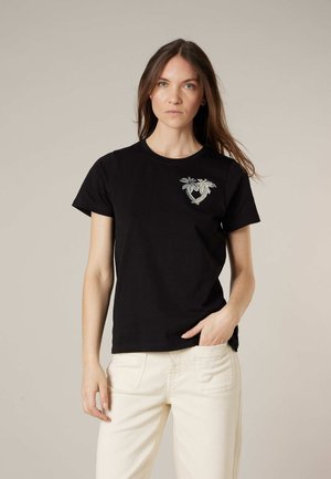 Femme portant un t-shirt noir à manches courtes avec un petit motif floral argenté sur le côté gauche de la poitrine, assorti à un pantalon blanc cassé, debout devant un fond uni.