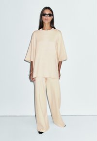 Beżowy, oversize'owy top z krótkimi rękawami w połączeniu z szerokimi beżowymi spodniami o luźnym kroju. Gładka faktura tkaniny; minimalistyczny design bez wzorów.