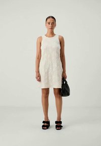 Balta bez piedurknēm kleita ar tekstūras detaļām, līdz ceļgaliem, kombinēta ar melnu somu un platformas sandālēm ar loka akcentiem.