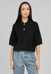 Pull polo tricoté noir à manches courtes, doté d'un ourlet côtelé et de trois boutons décoratifs sur le col. Porté avec un jean bleu clair.