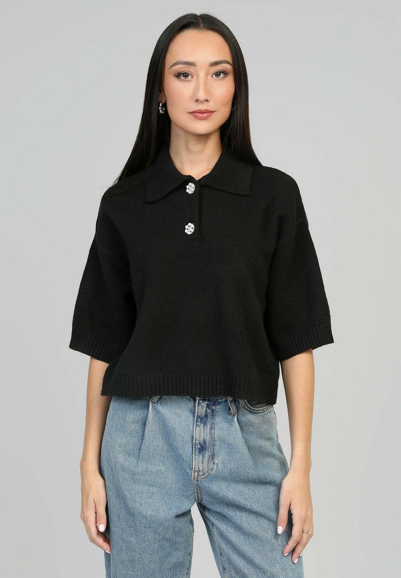 Pull polo tricoté noir à manches courtes, doté d'un ourlet côtelé et de trois boutons décoratifs sur le col. Porté avec un jean bleu clair.