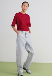 T-shirt rosso a maniche corte abbinato a jeans baggy azzurri con tasche laterali e dettagli cuciti. Il modello sta in piedi con le mani nelle tasche.