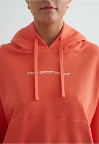 Koraalkleurige hoodie met trekkoorden, geribbelde textuur en een voorzak. Kenmerkt zich door witte borduring "AIM' SPORTSWEAR" op de borst.