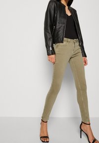 Jeans ajustados de color verde oliva, material de mezclilla, diseño ceñido, cintura media, con detalles de costura sutiles, combinados con una chaqueta de cuero negra y tacones.