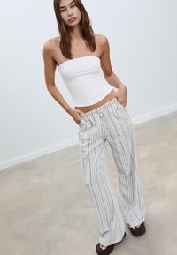 Bílý top bez ramínek v kombinaci se širokými kalhotami s šedými a bílými pruhy, které mají elastický pas a hladkou texturu s uvolněným střihem.