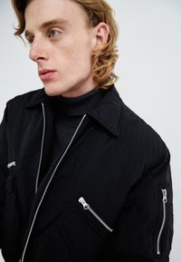 Chaqueta negra con un acabado texturizado, que cuenta con cremalleras plateadas en la parte frontal y en los bolsillos, además de un diseño con cuello, superpuesta sobre un negro de cuello alto.