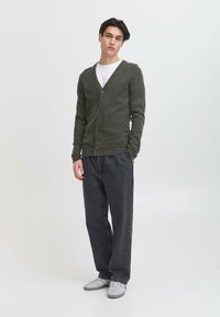 Mørkegrønn cardigan med V-hals, strukturert strikk og fem knapper, kombinert med løse svarte jeans og grå sneakers. Minimalistisk design.