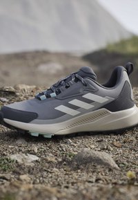 Chaussure de trail running grise et noire avec des bandes blanches et une semelle texturée sur un terrain rocailleux extérieur.