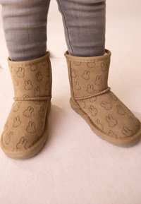 Hellbraune Wildlederstiefel mit einem flauschigen Inneren, das ein Muster aus kleinen Hasengesichtern zeigt. Kurze, abgerundete Form mit einer flexiblen Gummisohle.