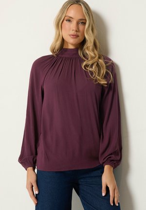 Vrouw met lang blond haar die een paarse blouse met hoge hals en lange mouwen en donkerblauwe broek draagt, staand tegen een witte achtergrond.