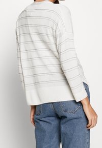 Lättstickad tröja i krämfärg med grå horisontella ränder och silvermetalliska detaljer. Har långa ärmar och en avslappnad passform.