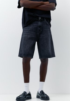 Személy fekete farmer térd hosszúságú rövidnadrágot, fehér zoknit és fekete bőr mokaszint visel, karját keresztbe téve áll egy egyszerű háttér előtt.