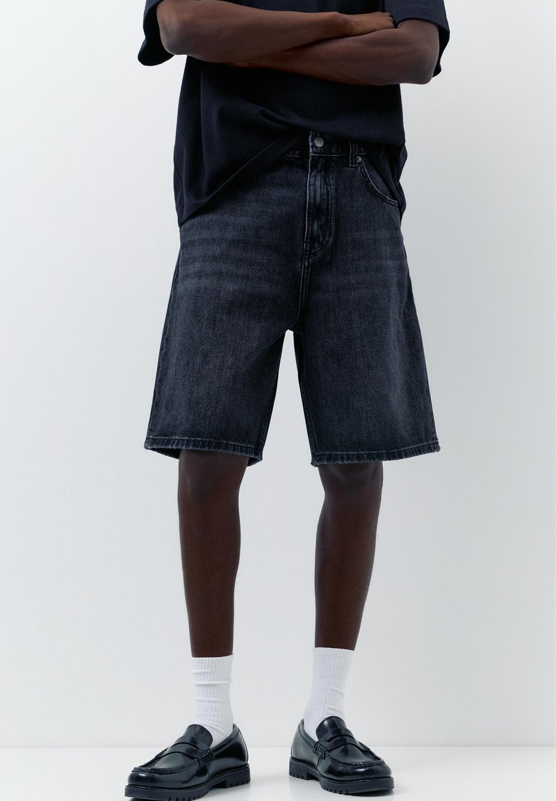 Personne portant un short en jean noir jusqu'aux genoux, des chaussettes blanches et des mocassins en cuir noirs, debout les bras croisés devant un fond uni.