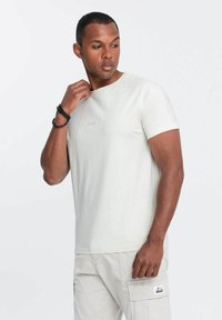 Camiseta de manga corta color beige claro con un cuello redondo y una textura sutil. Presenta un pequeño logo bordado en el pecho.