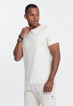 Camiseta de manga corta color beige claro con un cuello redondo y una textura sutil. Presenta un pequeño logo bordado en el pecho.